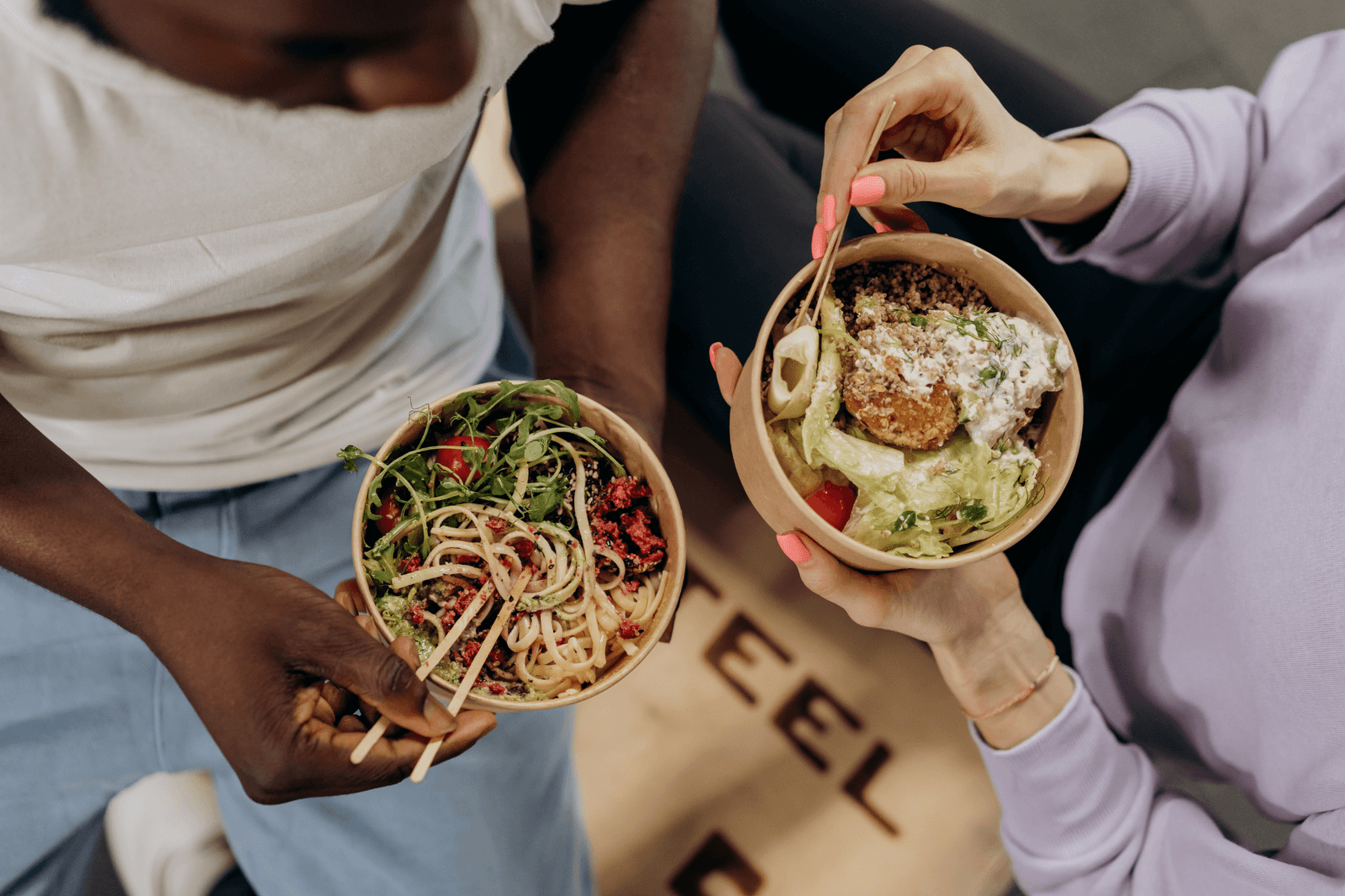 Duas pessoas segurando tigelas de comida saudável. Uma mão masculina segura uma tigela com macarrão, verduras e tomates, e uma mão feminina segura uma tigela com salada, um hambúrguer vegetariano e molho branco. O foco está nas tigelas, com o fundo intencionalmente fora de foco. A imagem representa a nutrição comportamental.