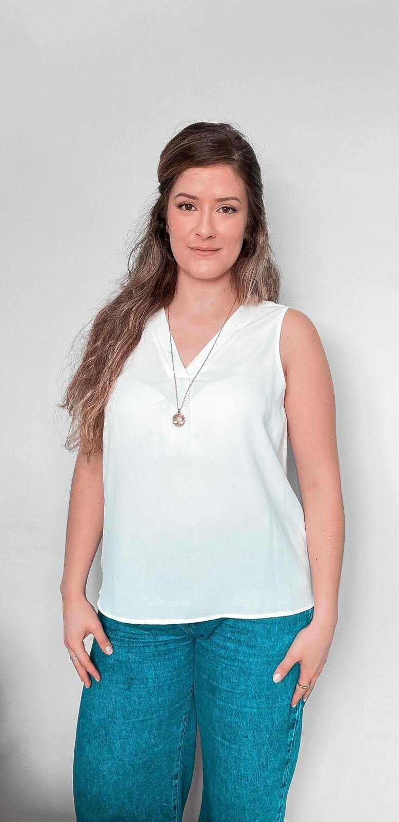 Karina Pankievicz, nutricionista em Curitiba, em pé, usando uma blusa branca sem mangas, um colar dourado e calça jeans, ela tem uma expressão serena e está olhando diretamente para a câmera. O fundo é neutro e simples, colocando o foco inteiramente nela.