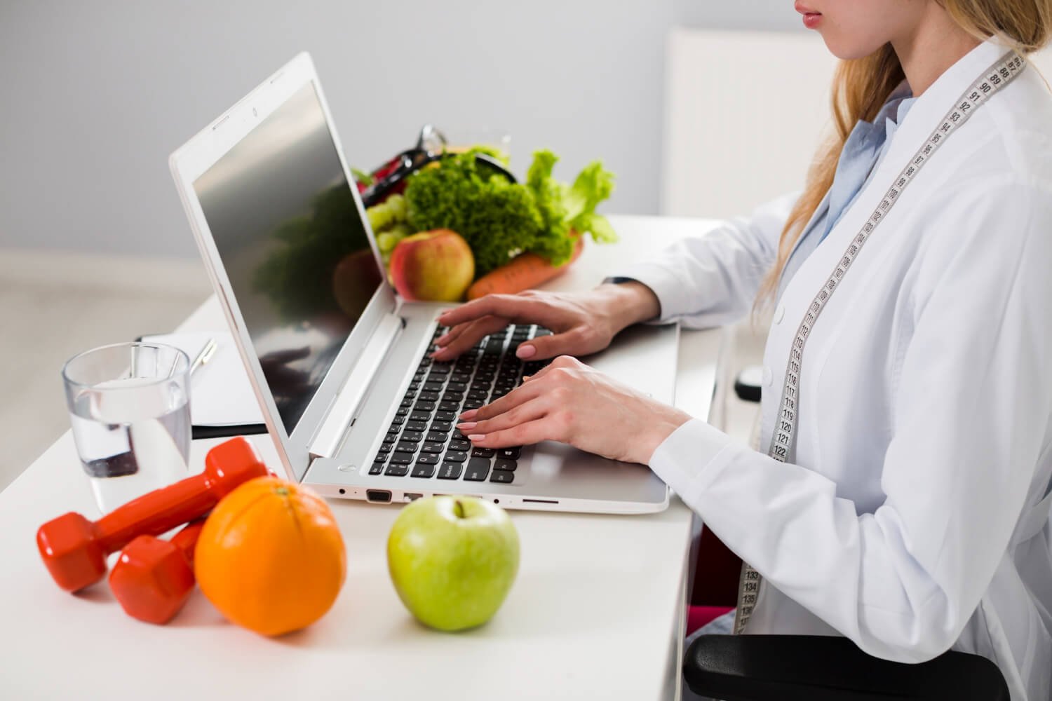 Uma nutricionista online está trabalhando em seu laptop, rodeada por frutas frescas como maçãs e uma laranja, e vegetais como alface e cenouras, indicando uma sessão de planejamento de dieta ou consulta nutricional. Ao lado do computador, há um copo de água e halteres vermelhos, sugerindo um foco no estilo de vida saudável que combina alimentação balanceada e exercícios físicos. A fita métrica sobre os ombros da nutricionista enfatiza a medição e o monitoramento do progresso do cliente.