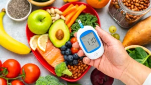 Pessoa segurando um glicosímetro que mostra leitura de 110 mg/dL, posicionada em frente a um prato em formato de coração com alimentos saudáveis para controle do diabetes, incluindo salmão, abacate, maçã verde, mirtilos, limão, brócolis, cenoura, tomate, banana, espinafre, grãos integrais e sementes de chia.